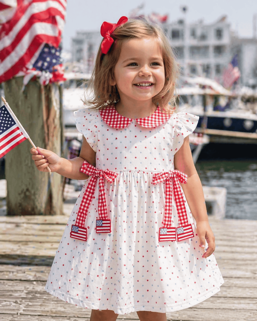 Young girl in a red, white, and blue dress holding an American flag on a dock.