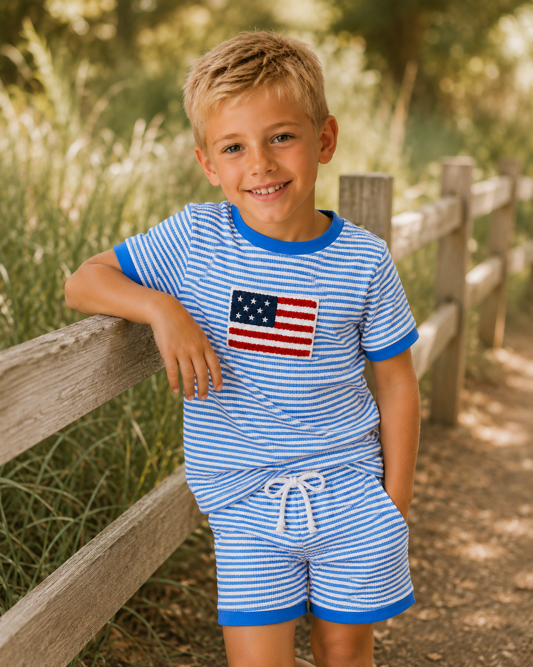 Young boy wearing a blue and white striped outfit with an American flag design, standing outdoors near a wooden fence.