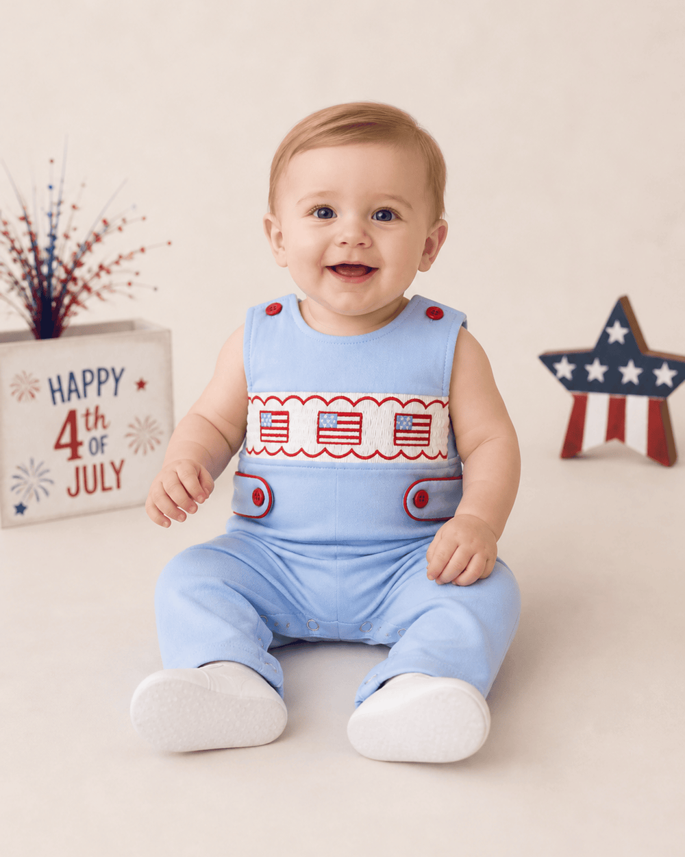 Baby wearing a blue outfit with American flag design, sitting on a white surface with 'Happy 4th of July' sign in the background.