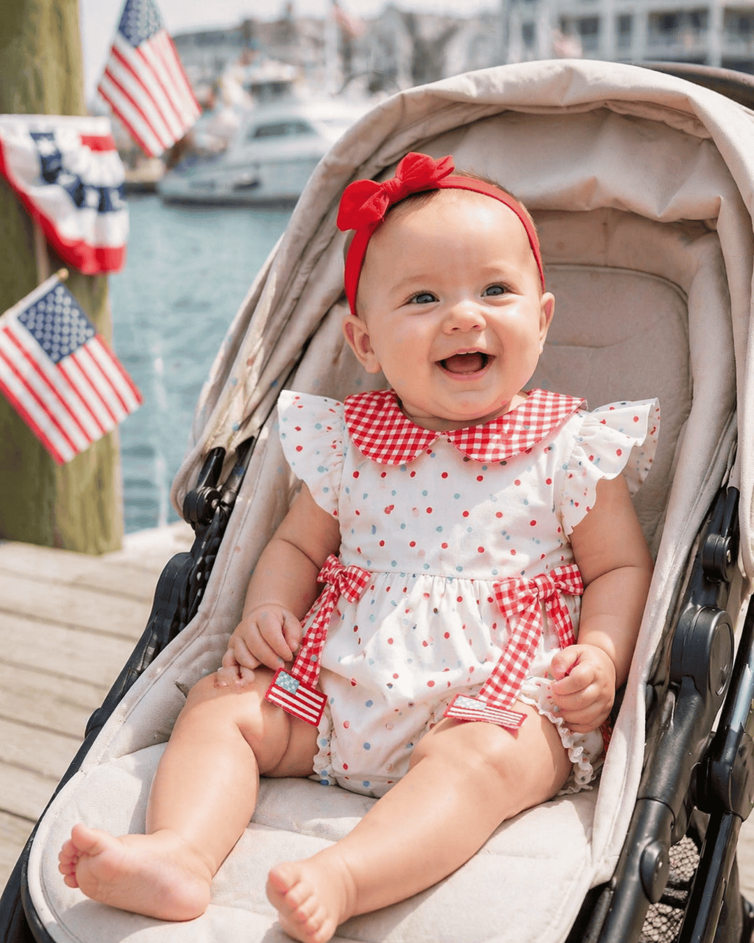 Baby in a stroller wearing a red and white outfit with a bow, with American flags in the background.