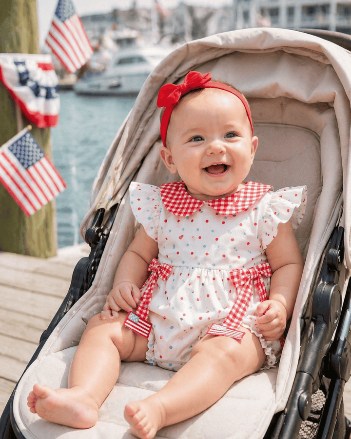 Baby in a stroller wearing a red and white outfit with a bow, with American flags in the background.
