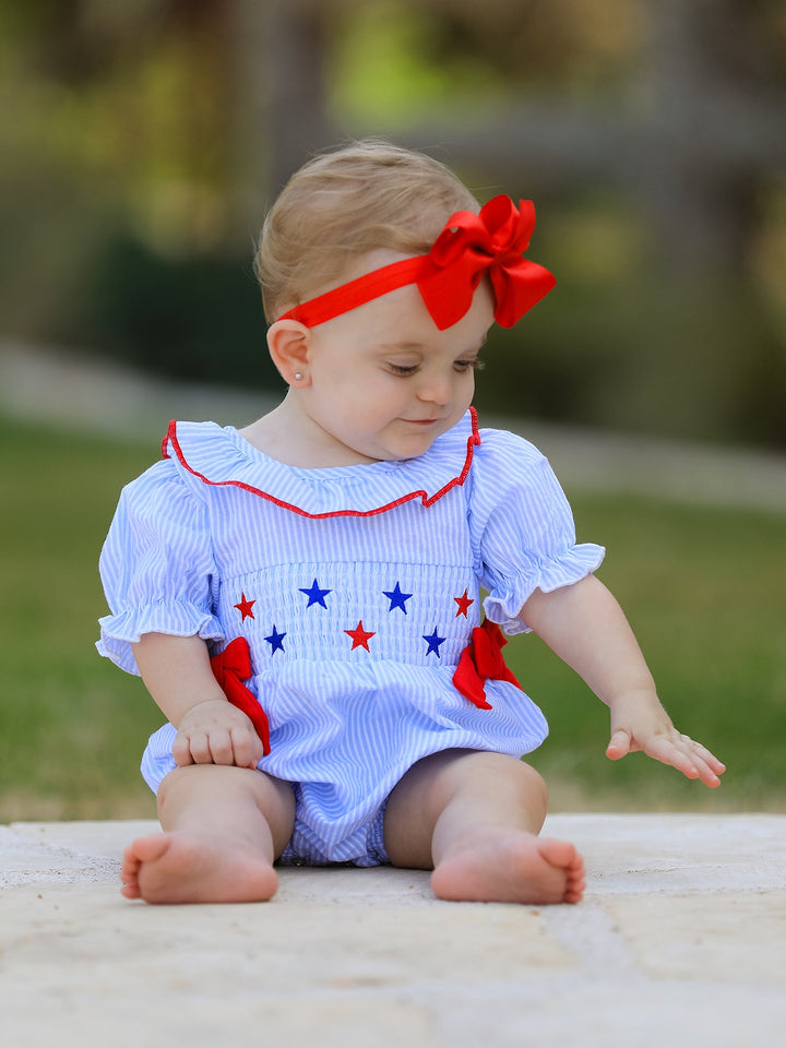 Baby wearing a blue outfit with red and white accents, sitting outdoors.