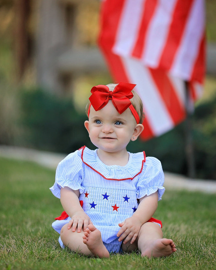 Baby in a patriotic outfit with a red bow sitting on grass in front of an American flag.