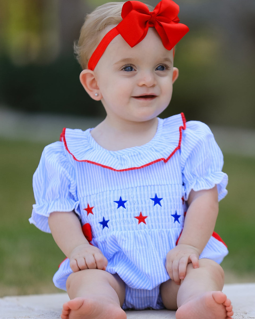 Baby wearing a white outfit with red and blue accents and a red bow headband.