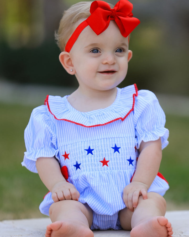 Baby wearing a white outfit with red and blue accents and a red bow headband.