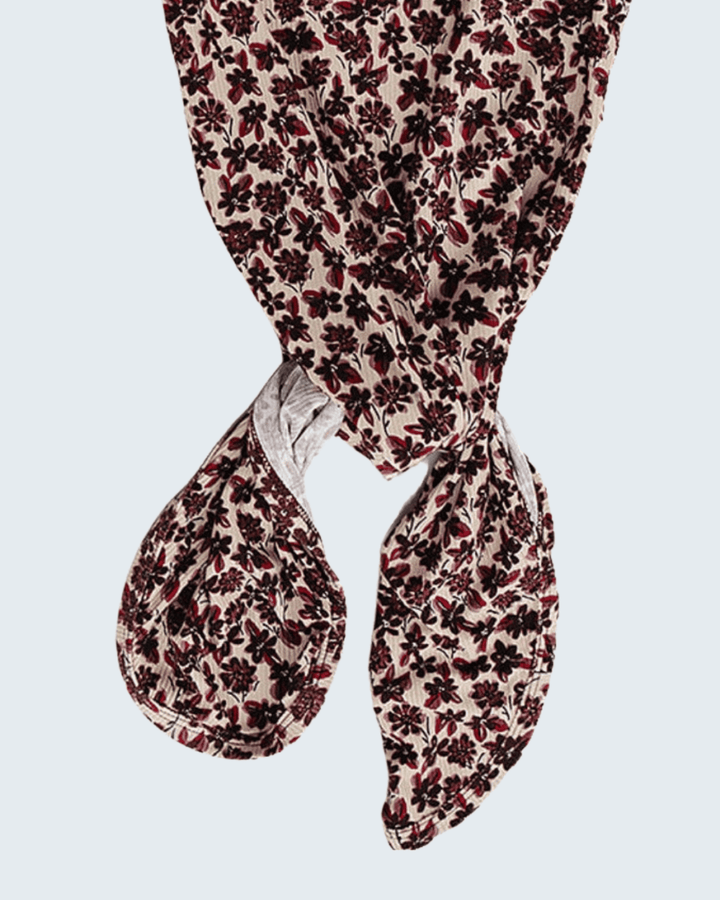 A knotted scarf featuring a dense floral pattern in dark red and black on a light beige background, shown against a solid light gray background.