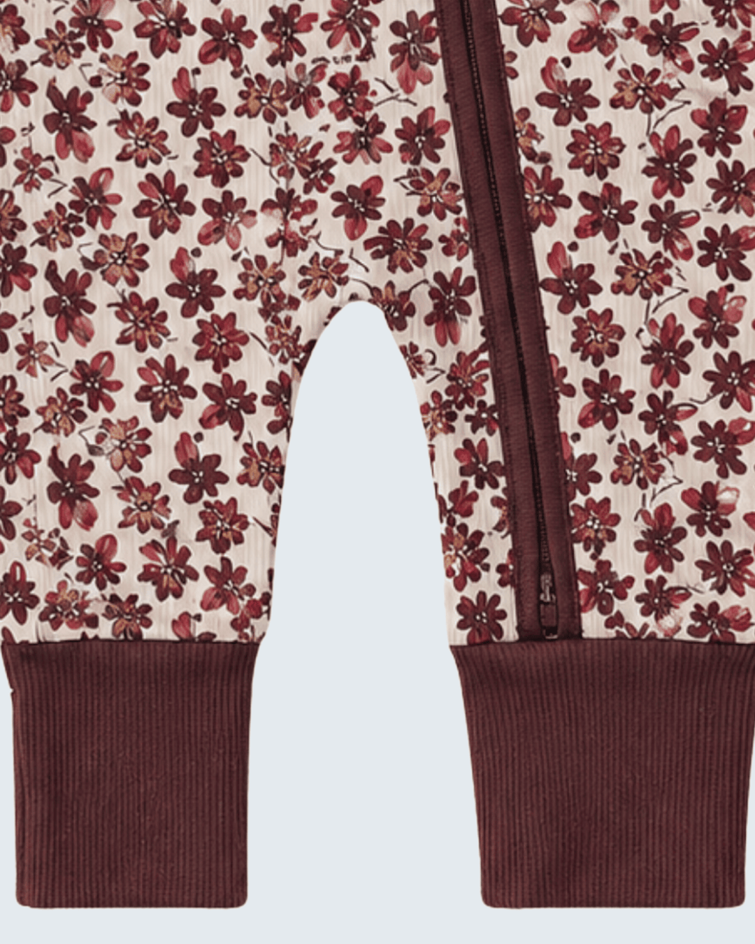Close-up of a pair of baby pants with an allover floral print in burgundy and dark red on a cream background, finished with solid burgundy ribbed cuffs and a matching zipper.