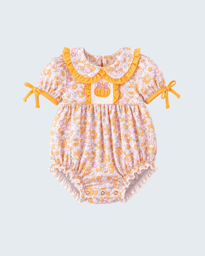 A baby romper with a yellow and pink floral pattern, featuring a ruffled yellow collar, a pumpkin applique at the center chest, and tie details on the sleeves, shown against a light gray background.