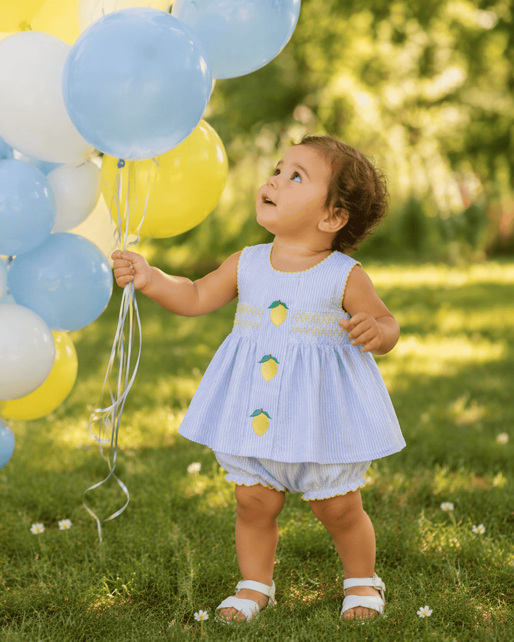 Child in a light blue dress with lemon designs holding balloons outdoors.