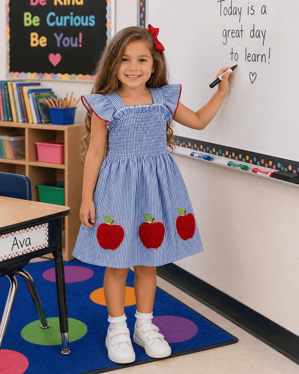Young girl in a classroom wearing a dress with red apples, standing next to a chalkboard.