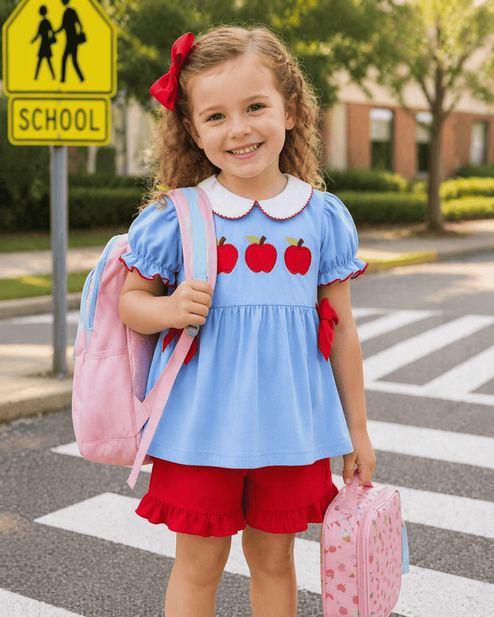 Young girl in a blue dress with red apples and red shorts standing on a crosswalk near a school sign.