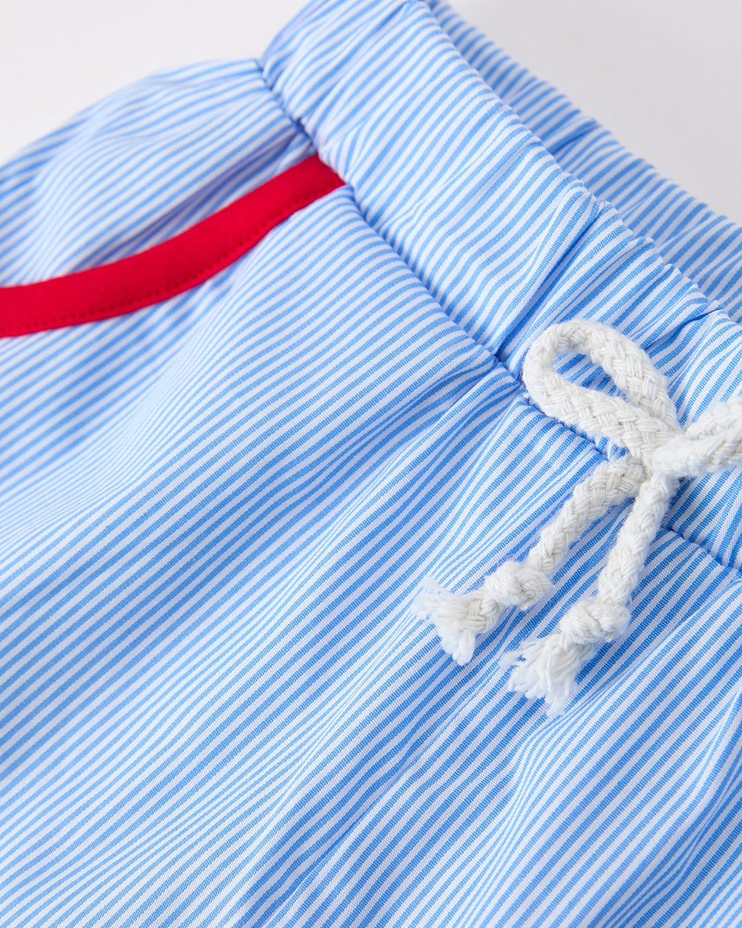 Close-up of blue striped shorts with a red waistband and white drawstring on a light gray background.