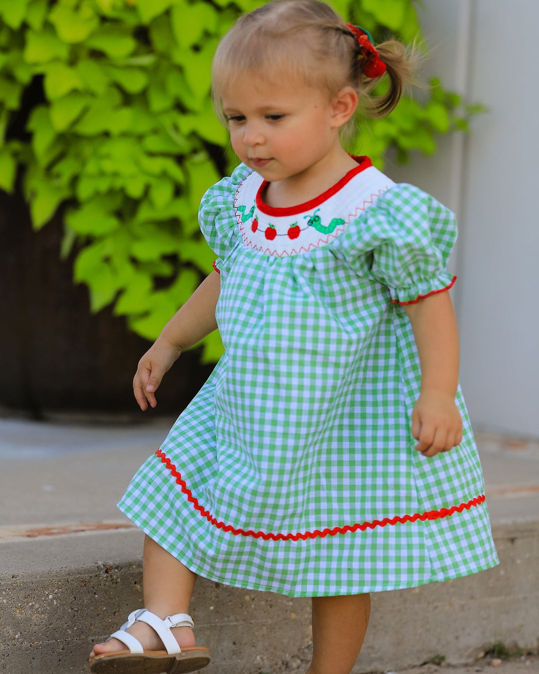 Young girl in a green checkered dress with red trim standing outdoors.