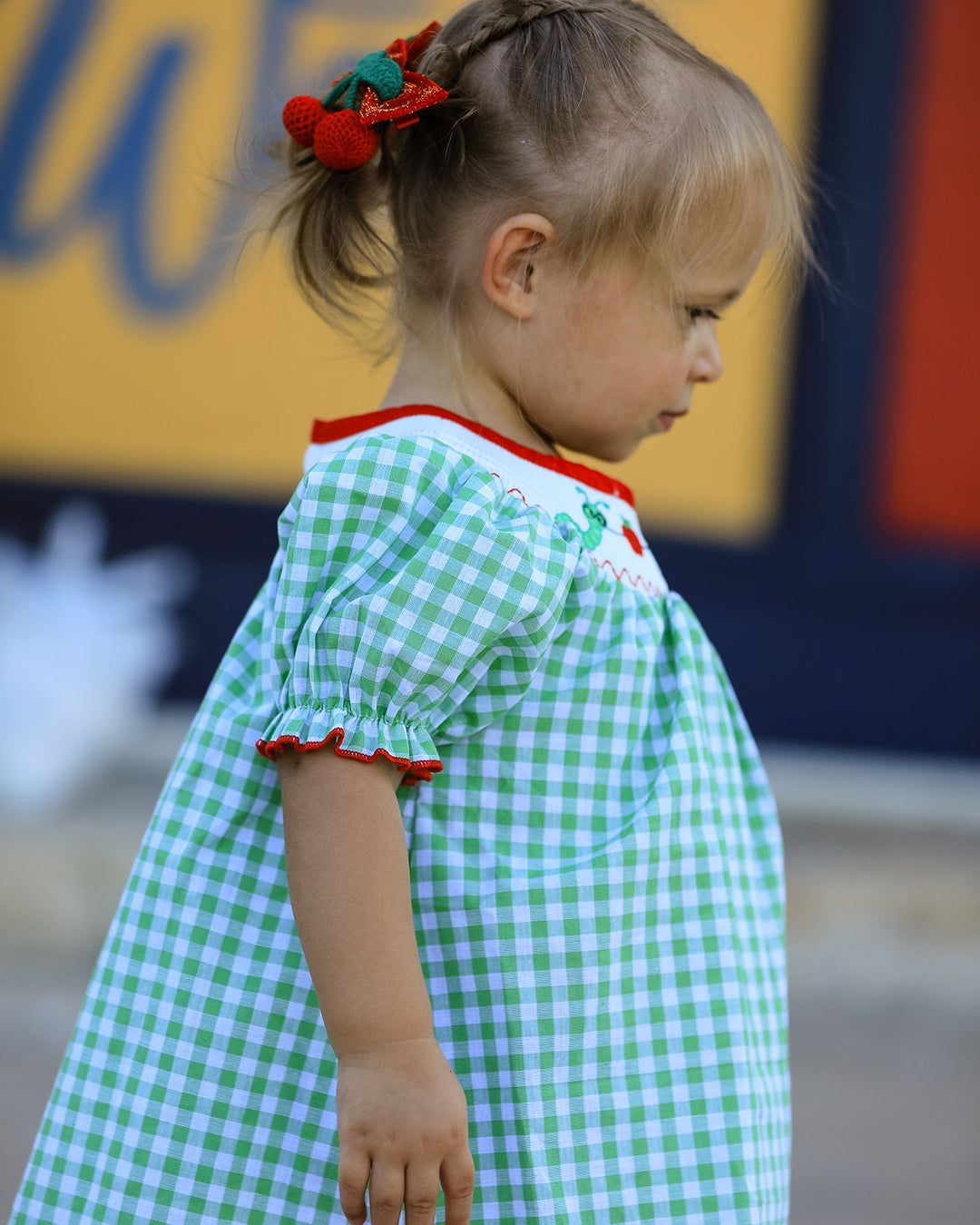 Child wearing a green checkered dress with red trim, standing outdoors.
