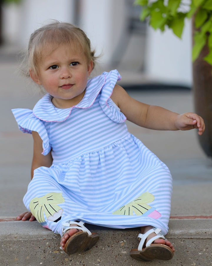 Child wearing a blue dress with ruffled sleeves and leaf designs, sitting on a concrete surface.