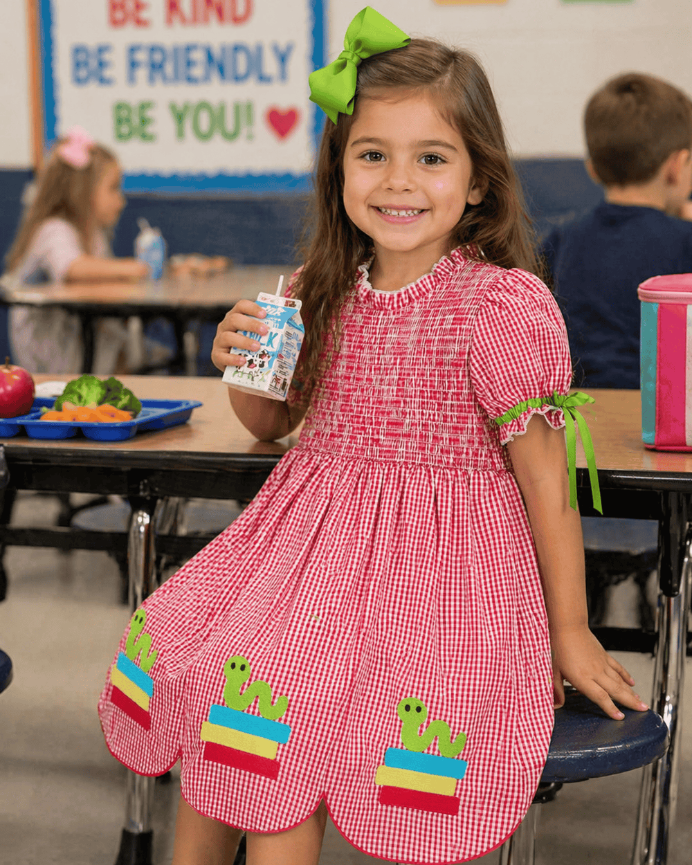 Young girl in a classroom wearing a red checkered dress with snake designs, holding a juice box.