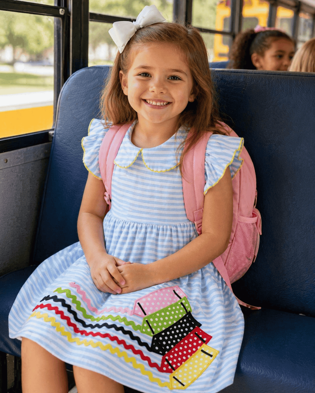 Young girl with a pink backpack sitting on a bus, wearing a blue dress with colorful details.