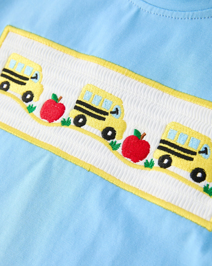 Light blue fabric with embroidered yellow school buses and red apples on a white border.