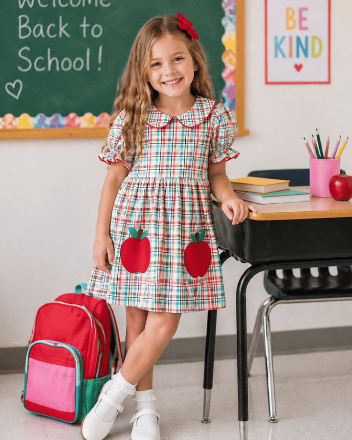 Young girl in a plaid dress with apple designs, standing in a classroom setting.