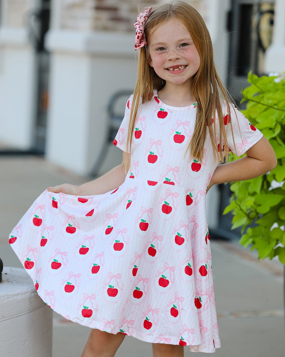 Young girl wearing a white dress with red apple patterns outdoors.