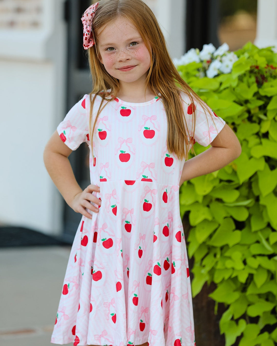 Young girl wearing a white dress with red apple pattern outdoors.
