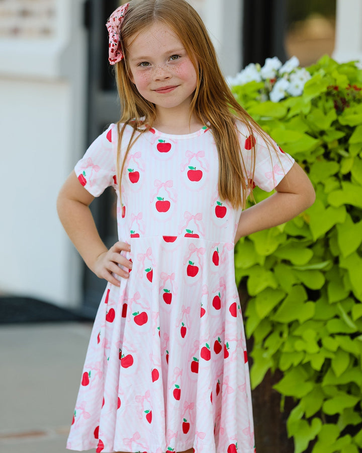 Young girl wearing a white dress with red apple pattern outdoors.