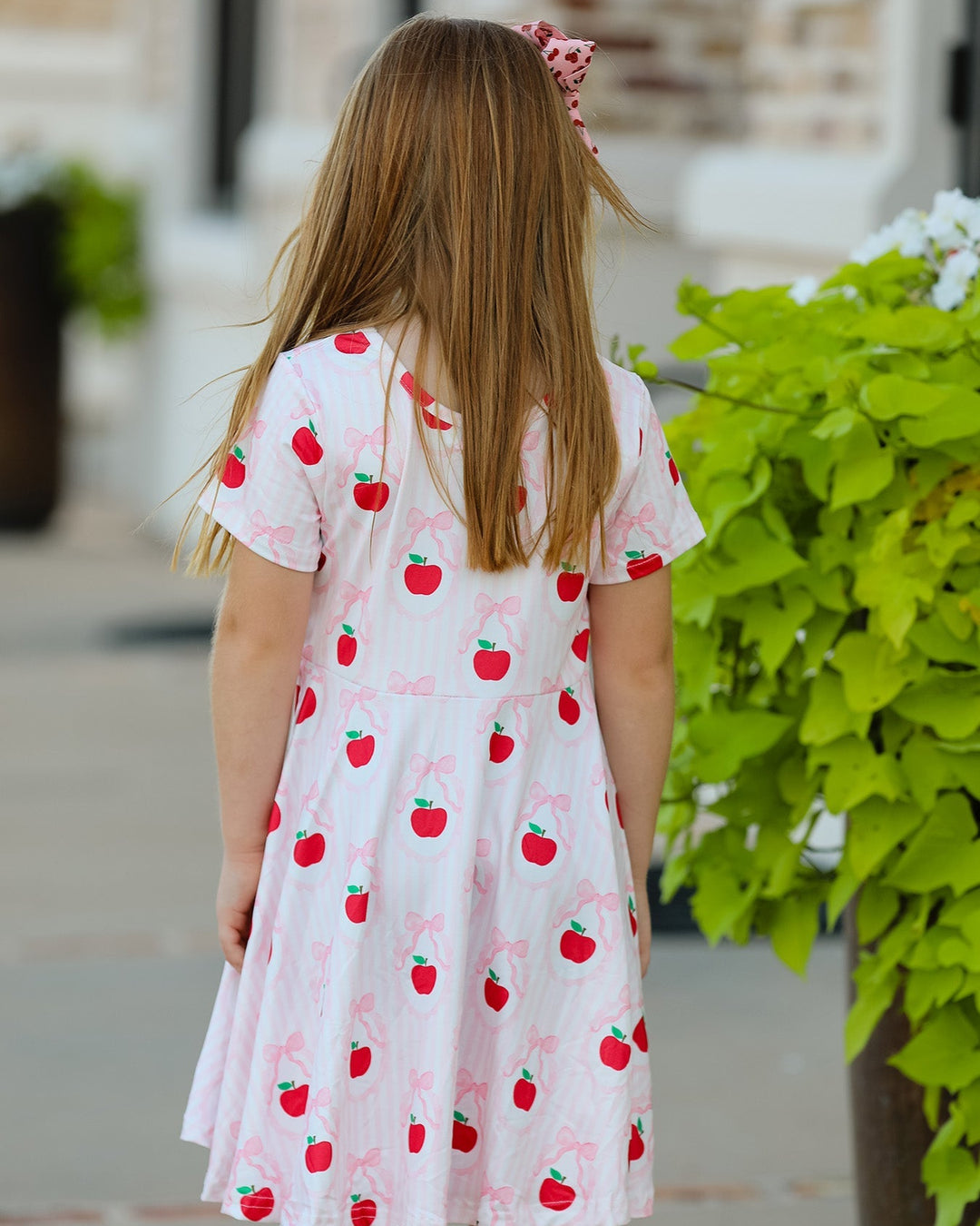 Young girl wearing a white dress with red apple patterns, standing outdoors.