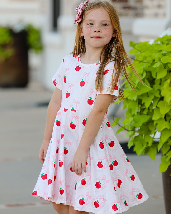 Young girl wearing a white dress with red apple patterns outdoors.