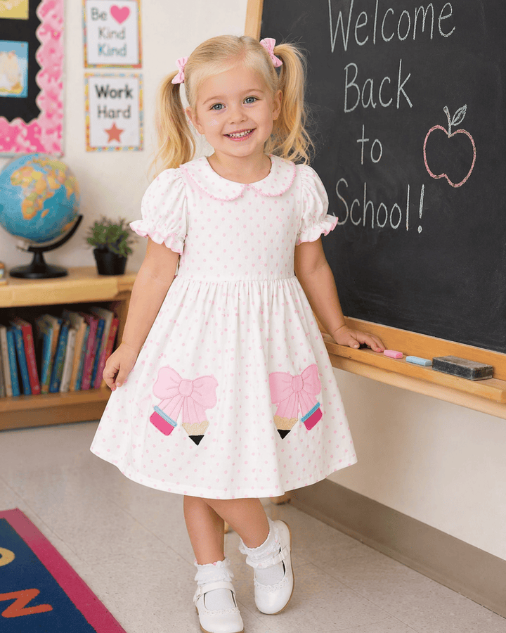 Young girl in a white dress with pink accents standing in front of a blackboard in a classroom setting.