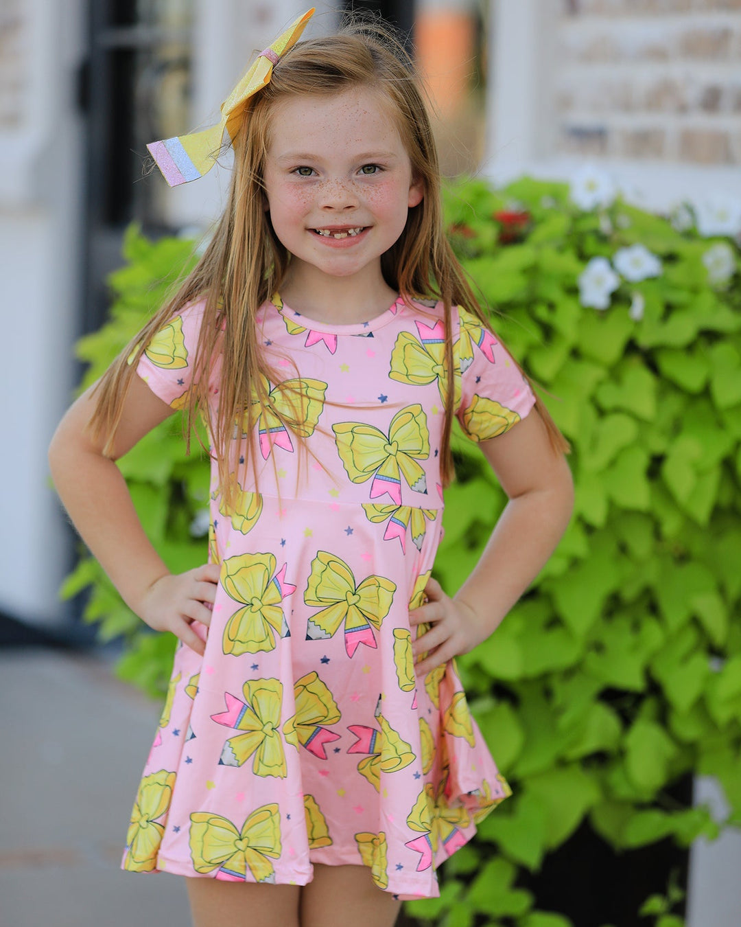 Young girl wearing a pink dress with yellow bow pattern, standing outdoors with greenery in the background.