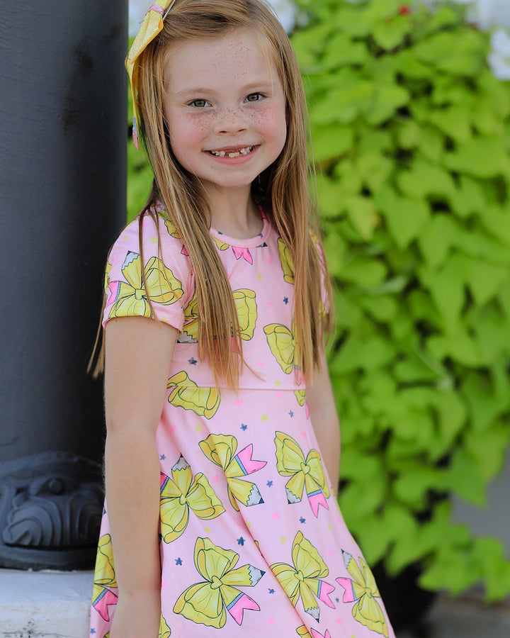Young girl wearing a pink dress with yellow bow pattern, standing outdoors.