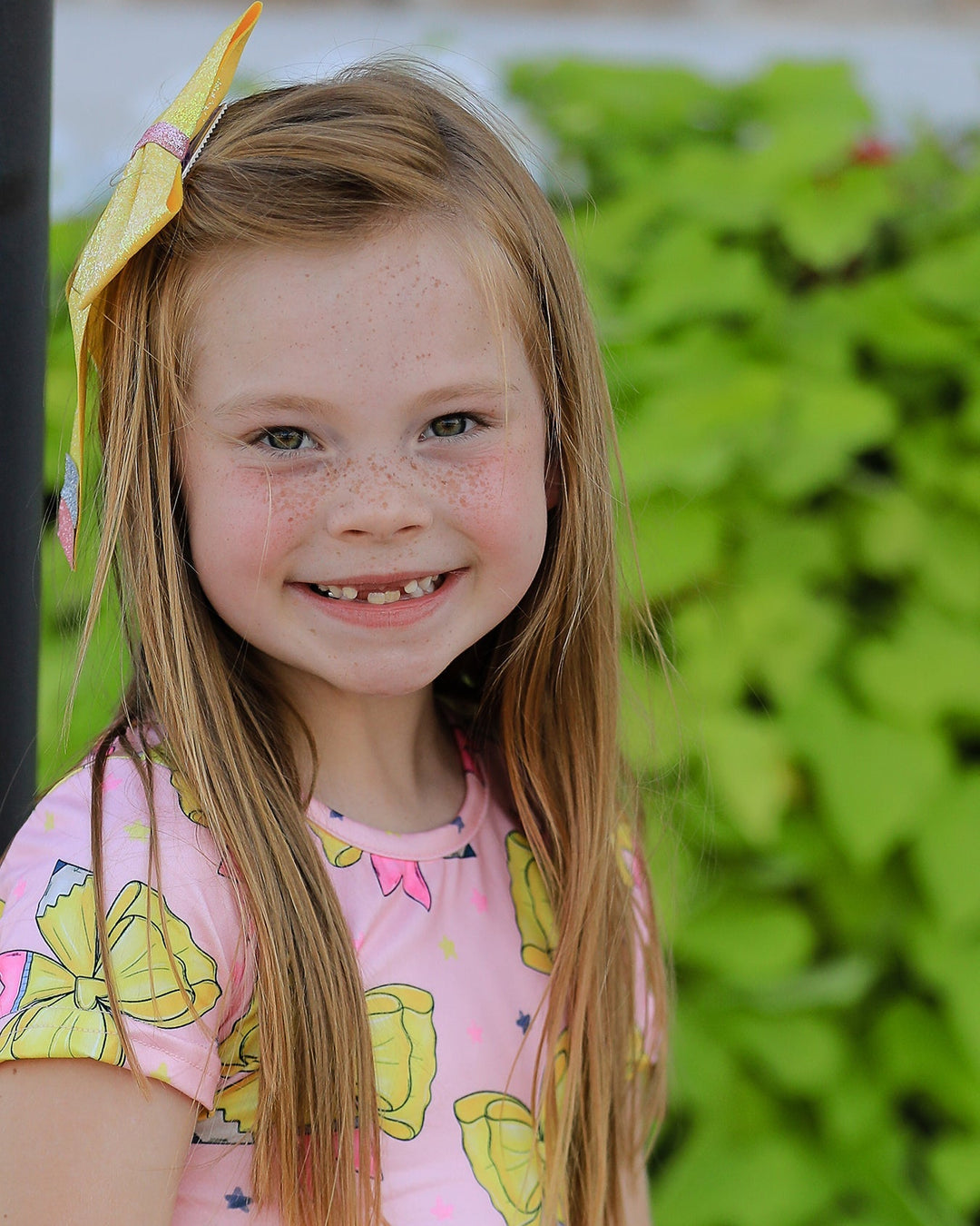 Young girl with a yellow bow in her hair, wearing a pink shirt with yellow graphics, standing against a green leafy background.