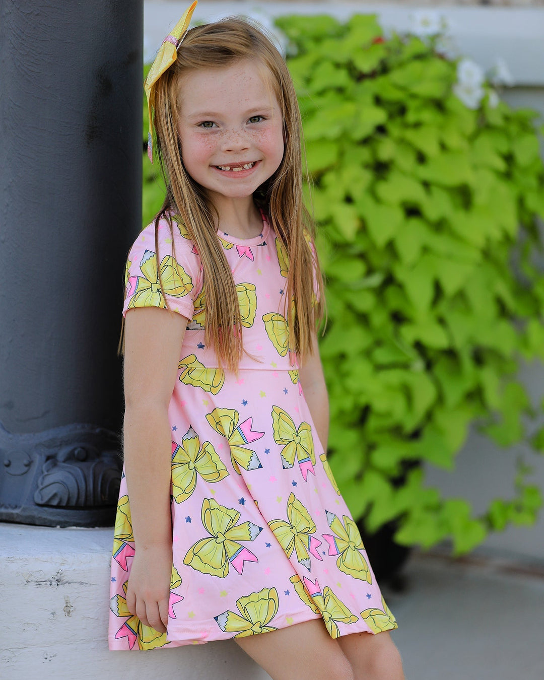 Young girl wearing a pink dress with yellow bow pattern, standing outdoors.