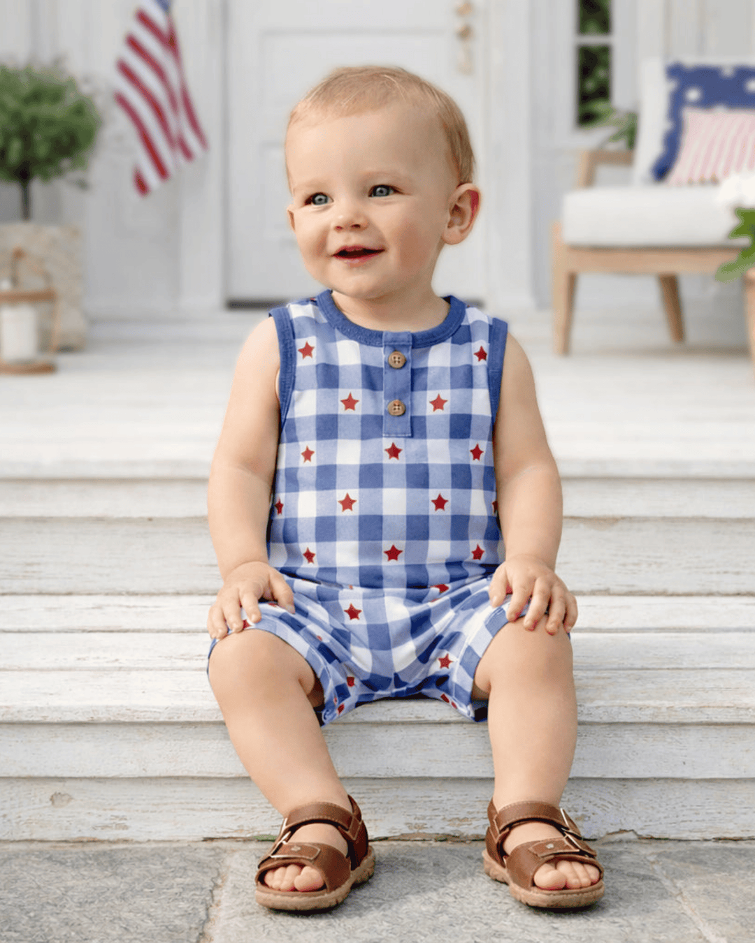 Child wearing a blue checkered romper with red stars on a wooden porch.