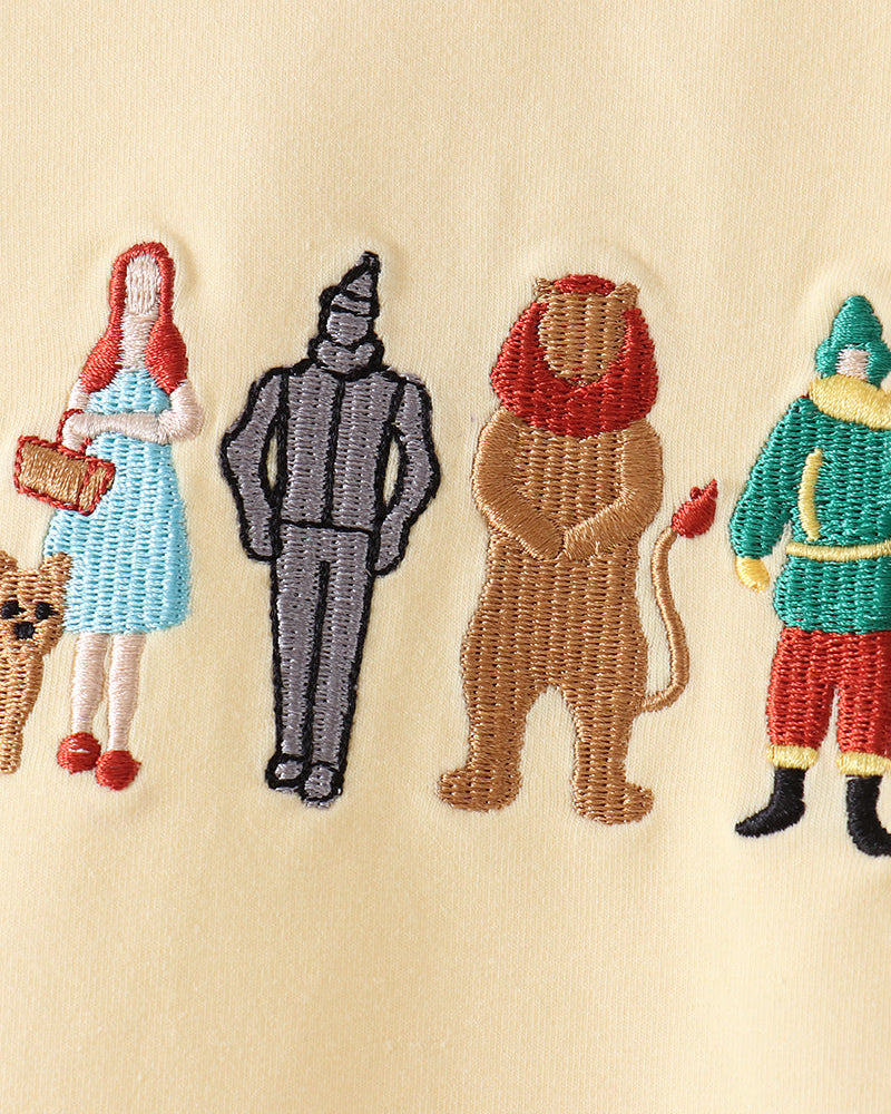Close-up of embroidered characters on a cream fabric background, including a woman in a blue dress, a gray wizard, a lion with a red mane, and a green-clad figure.