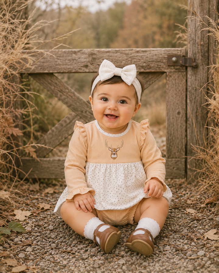 A baby girl sitting outdoors in front of a rustic wooden gate, wearing a peach and white polka dot romper, a white bow headband, and brown shoes.