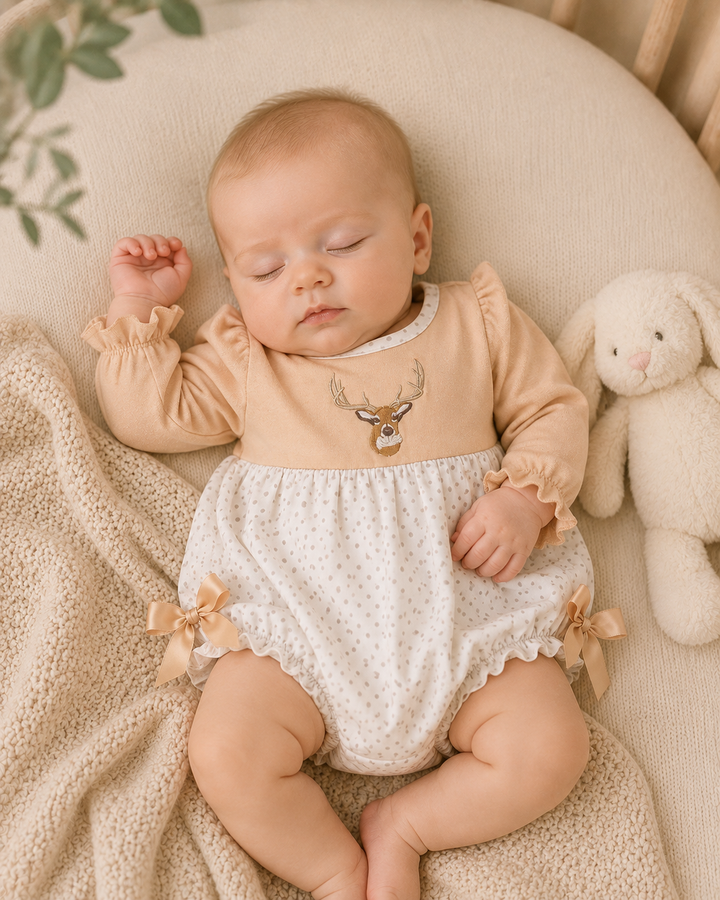 A sleeping baby wearing a beige long-sleeve romper with a deer graphic and white dotted bloomers, lying on a textured cream blanket next to a white stuffed bunny.