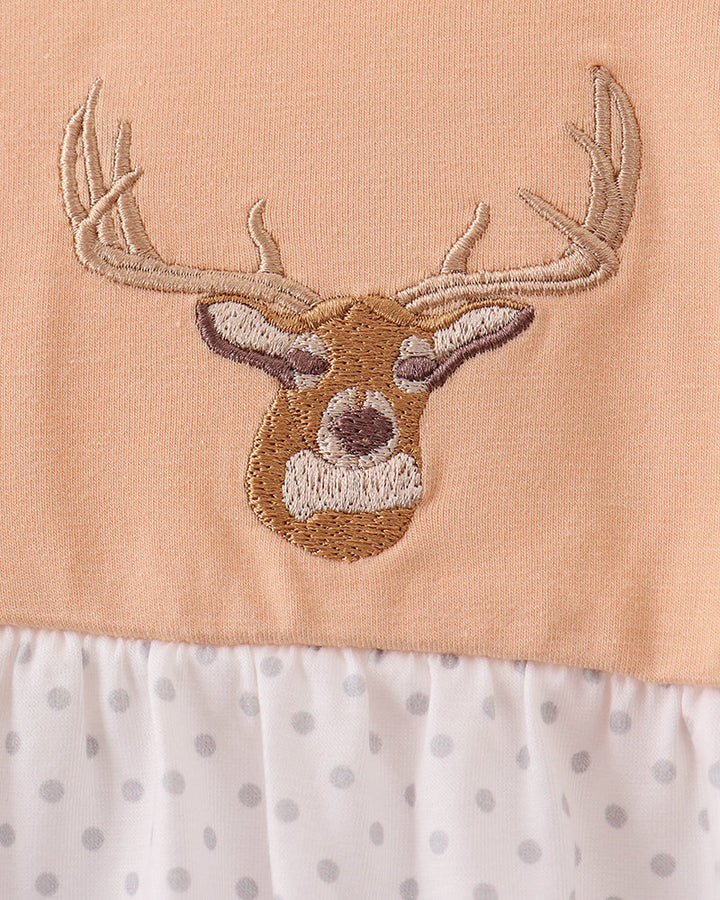 Close-up of a baby dress featuring a peach-colored top with an embroidered deer head motif, a white ruffled neckline with gray polka dots, and a white ruffled hem with gray polka dots.