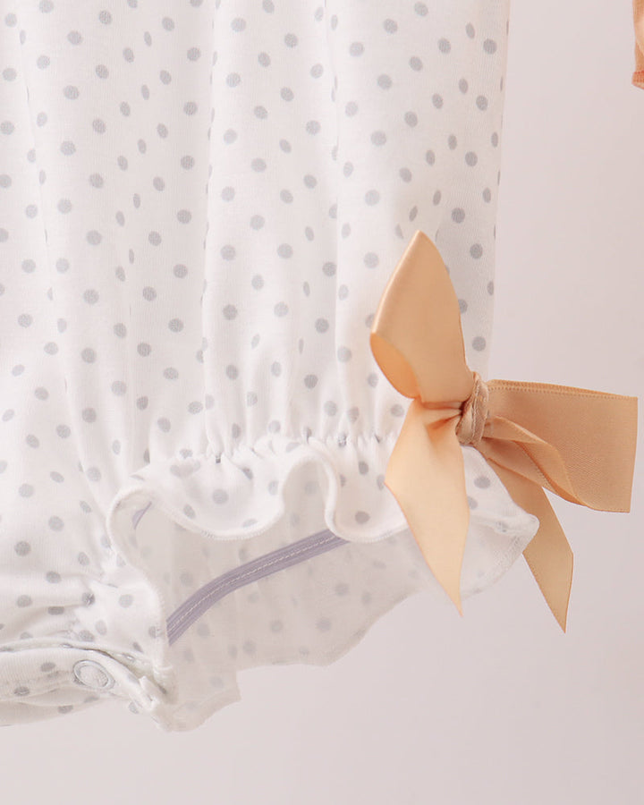 White baby bloomers with small gray polka dots and a peach-colored satin bow tied on the side, shown against a light pink background.