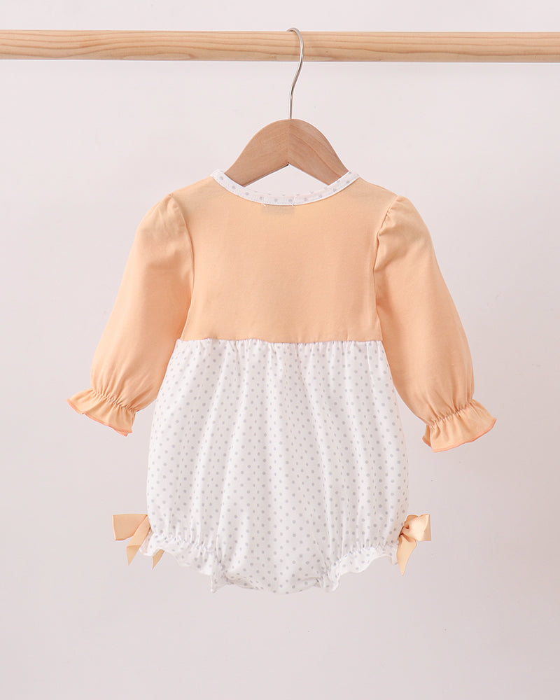 A baby romper hanging on a wooden hanger from a wooden clothes rack. The romper features a peach-colored top and sleeves with a white polka-dot skirt and lace trim.