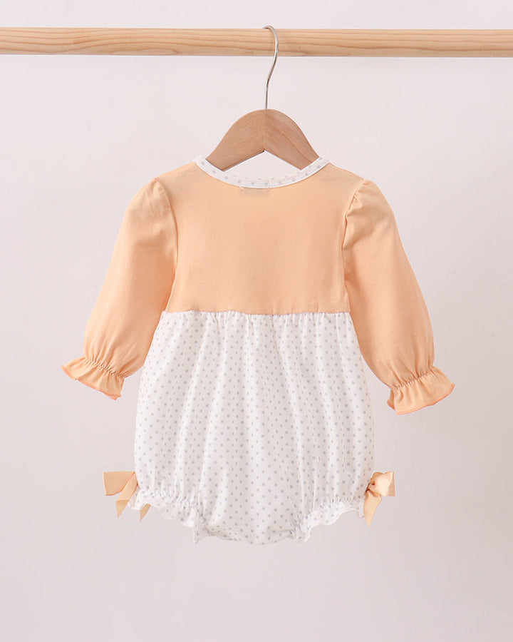 A baby romper hanging on a wooden hanger from a wooden clothes rack. The romper features a peach-colored top and sleeves with a white polka-dot skirt and lace trim.