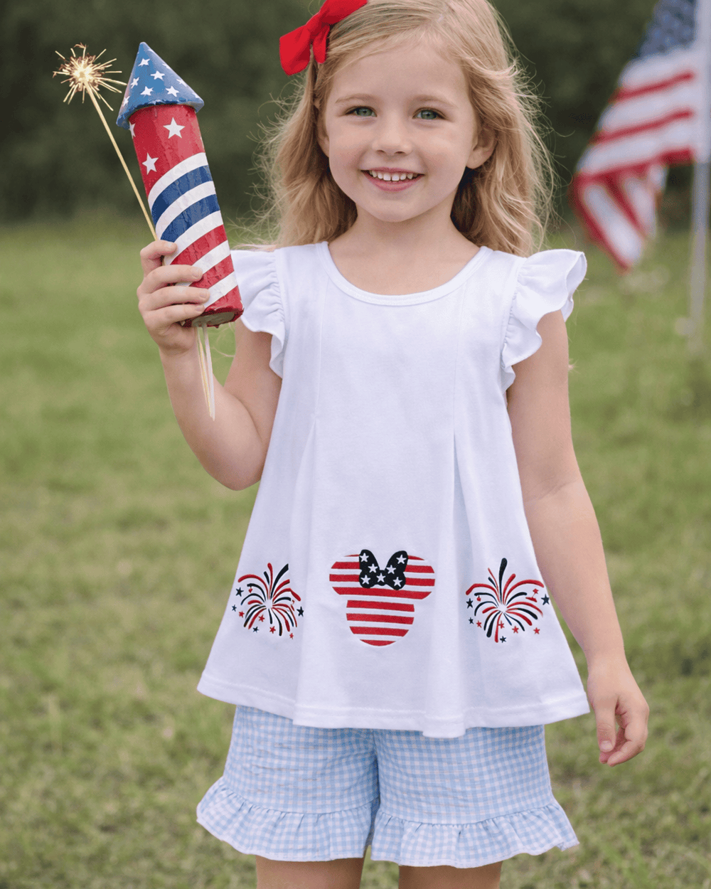 Young girl in a white top with patriotic designs and light blue shorts holding a sparkler outdoors.