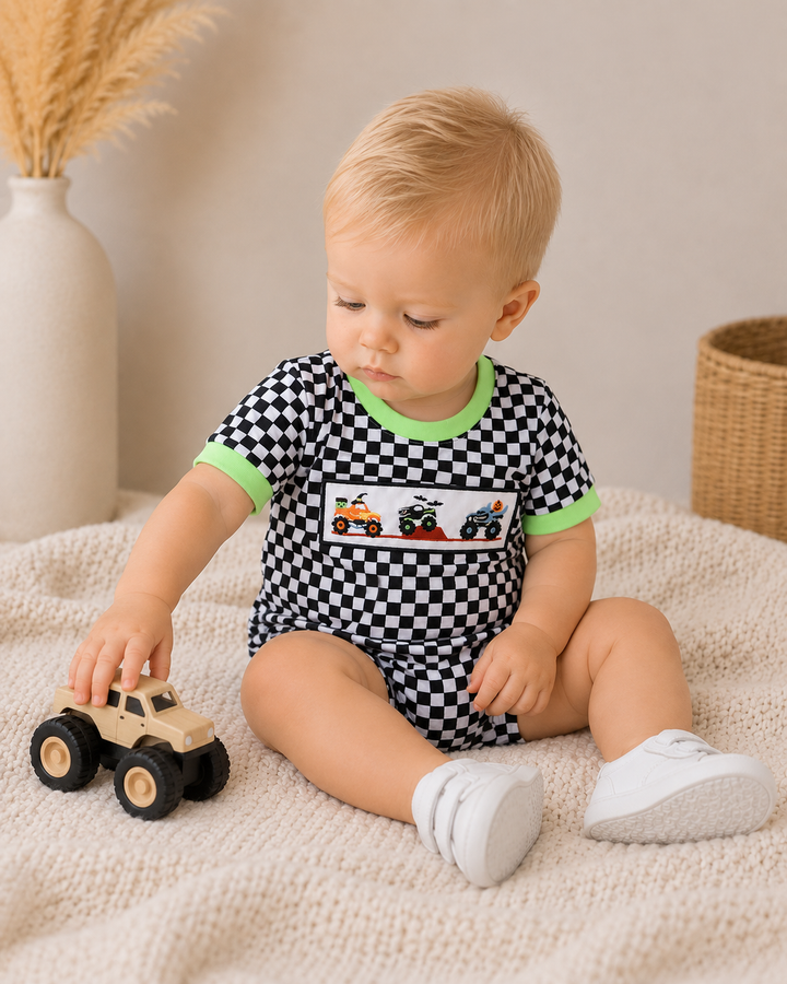 A baby sitting on a cream knit blanket wearing a black and white checkered short-sleeve romper with green trim and a graphic of race cars on the chest. The baby is holding a small toy truck.
