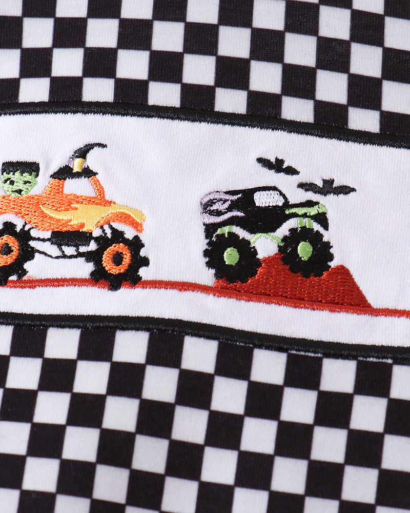 A close-up of a black and white checkered fabric with a horizontal embroidered band featuring a green monster truck, a black monster truck, a green-and-orange Halloween ATV, bats, and a red dirt mound.