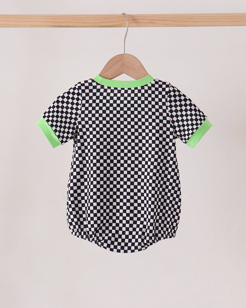 A short-sleeve baby romper with a black and white checkerboard pattern, featuring bright green trim on the collar and sleeve cuffs, hanging on a wooden hanger from a wooden clothes rack.