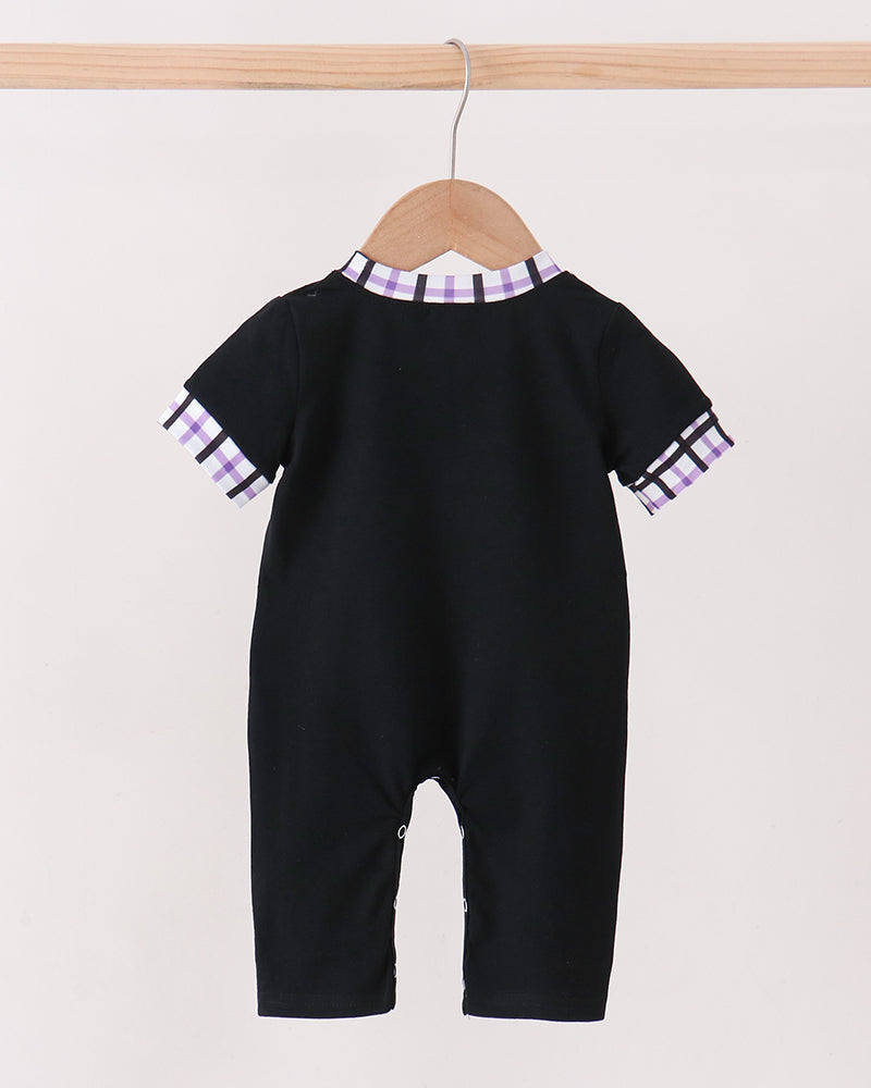 A baby short-sleeve black romper hanging on a wooden hanger from a light wood rod, featuring purple and white checkered trim at the collar and sleeve cuffs, set against a plain off-white background.