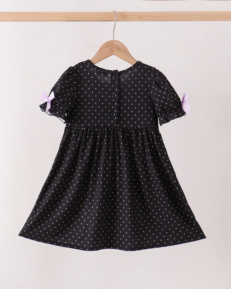 A black short-sleeve dress with white polka dots hanging on a wooden hanger against a plain light background. The dress has a gathered waist and small lavender bows on the sleeves.