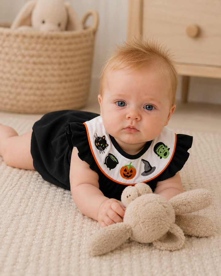 A baby lying on a cream carpet wearing a black romper and a white bib with Halloween-themed embroidered characters, holding a plush bunny toy.