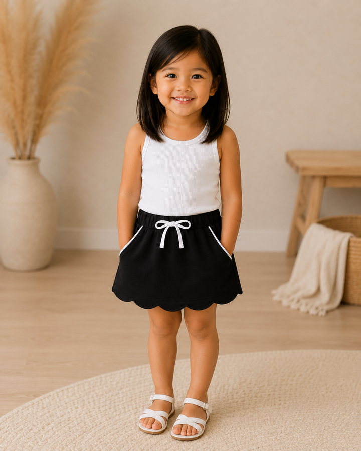 A young girl stands smiling in a bright indoor room, wearing a sleeveless white top and a black skirt with a white drawstring and white side trim. The background includes a beige wall, light wood floor, a vase with dried grass, a small wooden bench, and a woven throw blanket.