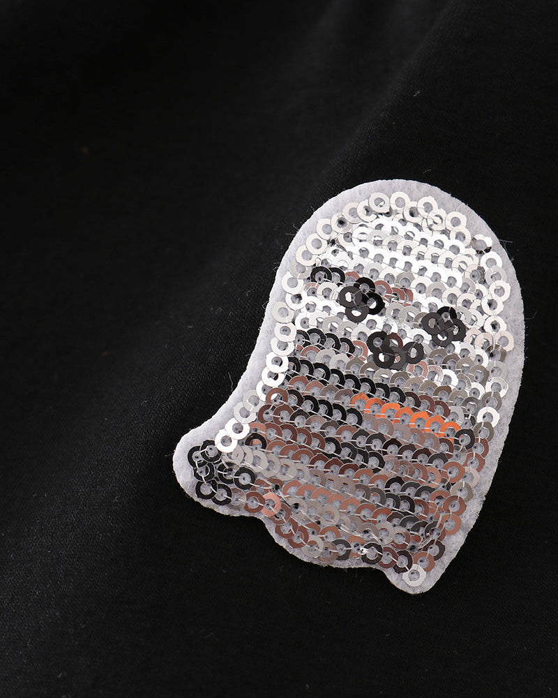 A ghost-shaped sequin patch with a white border, featuring black and silver sequins for a face and orange sequins for the body, displayed on a black fabric background.